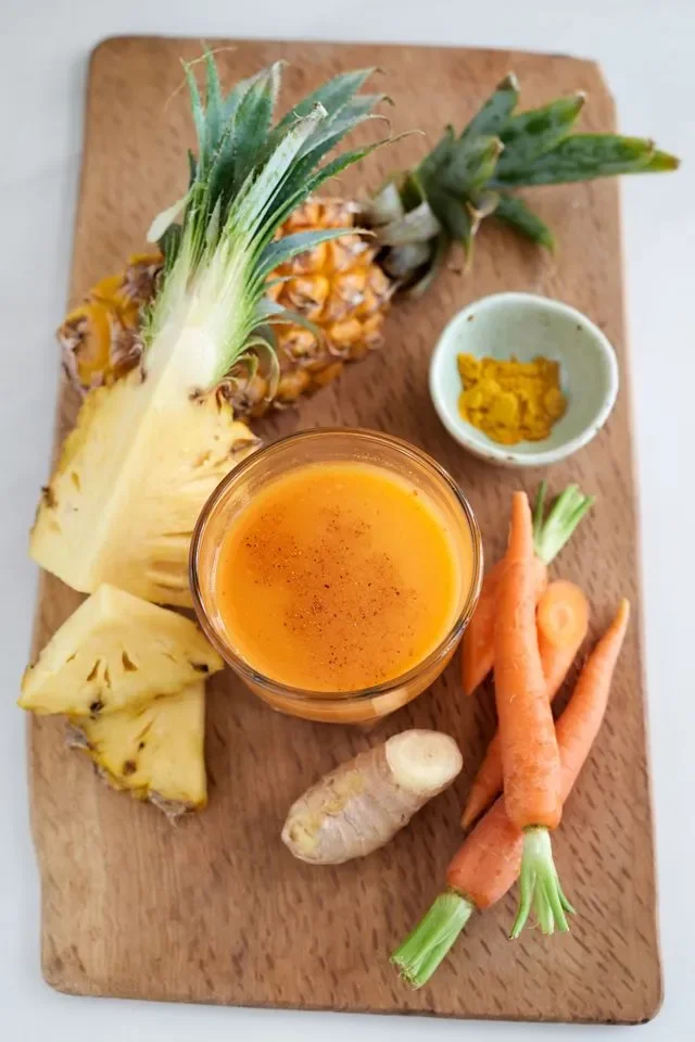 Glass of fresh juice made with pineapple, carrots, and ginger on a wooden board, surrounded by sliced pineapple, whole carrots, ginger root, and a small bowl of turmeric.