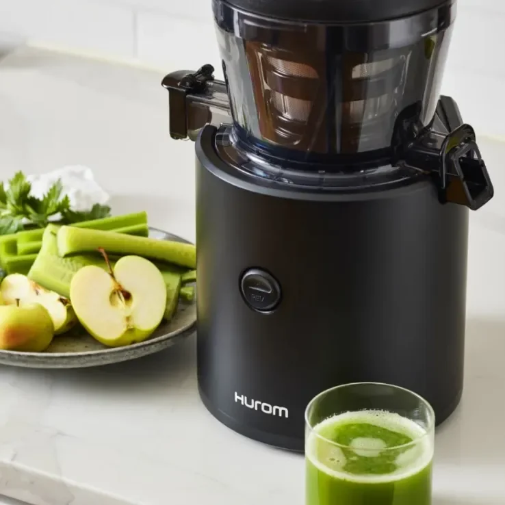 Hurom slow juicer on a kitchen counter with a glass of fresh green juice, alongside sliced apples, celery, and parsley ready for juicing.