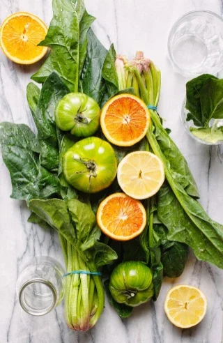 Top-down view of fresh ingredients including spinach, celery, green tomatoes, oranges, and lemon arranged on a marble surface with empty glasses nearby.