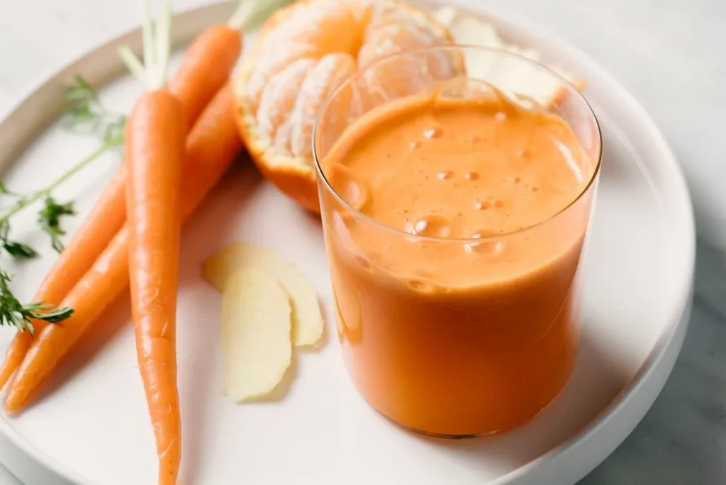 Glass of bright orange carrot juice served with whole carrots, peeled orange, and slices of fresh ginger on a plate.