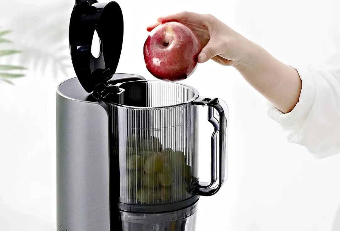 Hand placing a whole apple into the top of a Hurom slow juicer, with the lid open and fruit already inside the chamber.