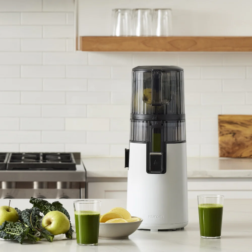 Hurom slow press juicer on a modern kitchen counter with fresh green juice, apples, kale, and lemon slices.