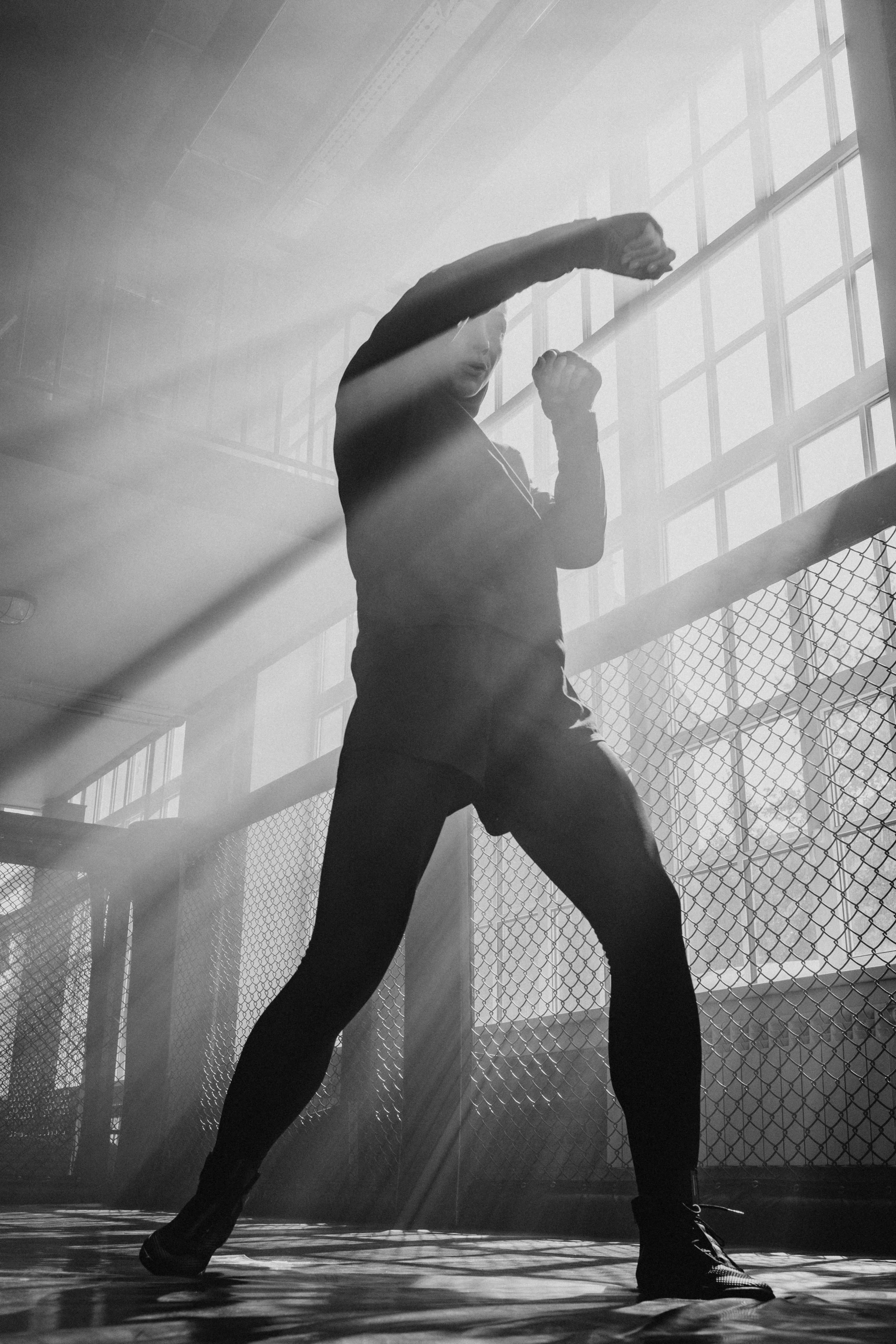 A person in athletic attire, including black leggings and sneakers, practicing boxing or martial arts in a gym with large windows and sunlight streaming through, casting shadows on the floor.