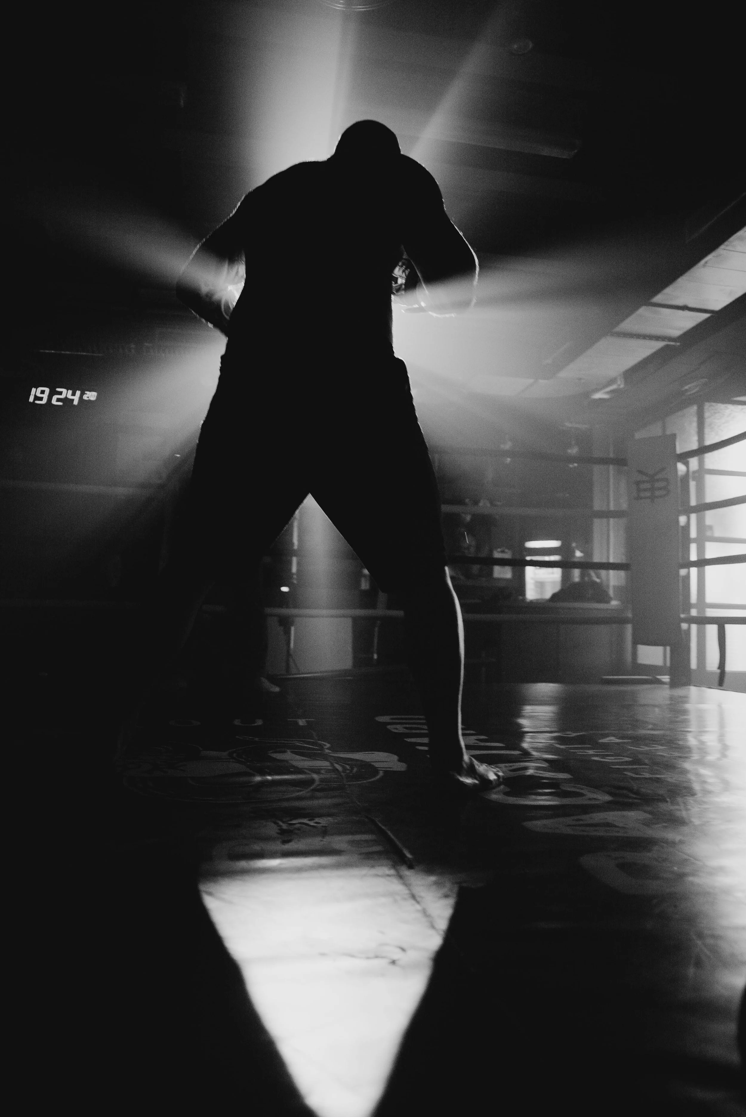 Silhouette of a person in a dimly lit boxing gym with boxing ring ropes visible on the right side.