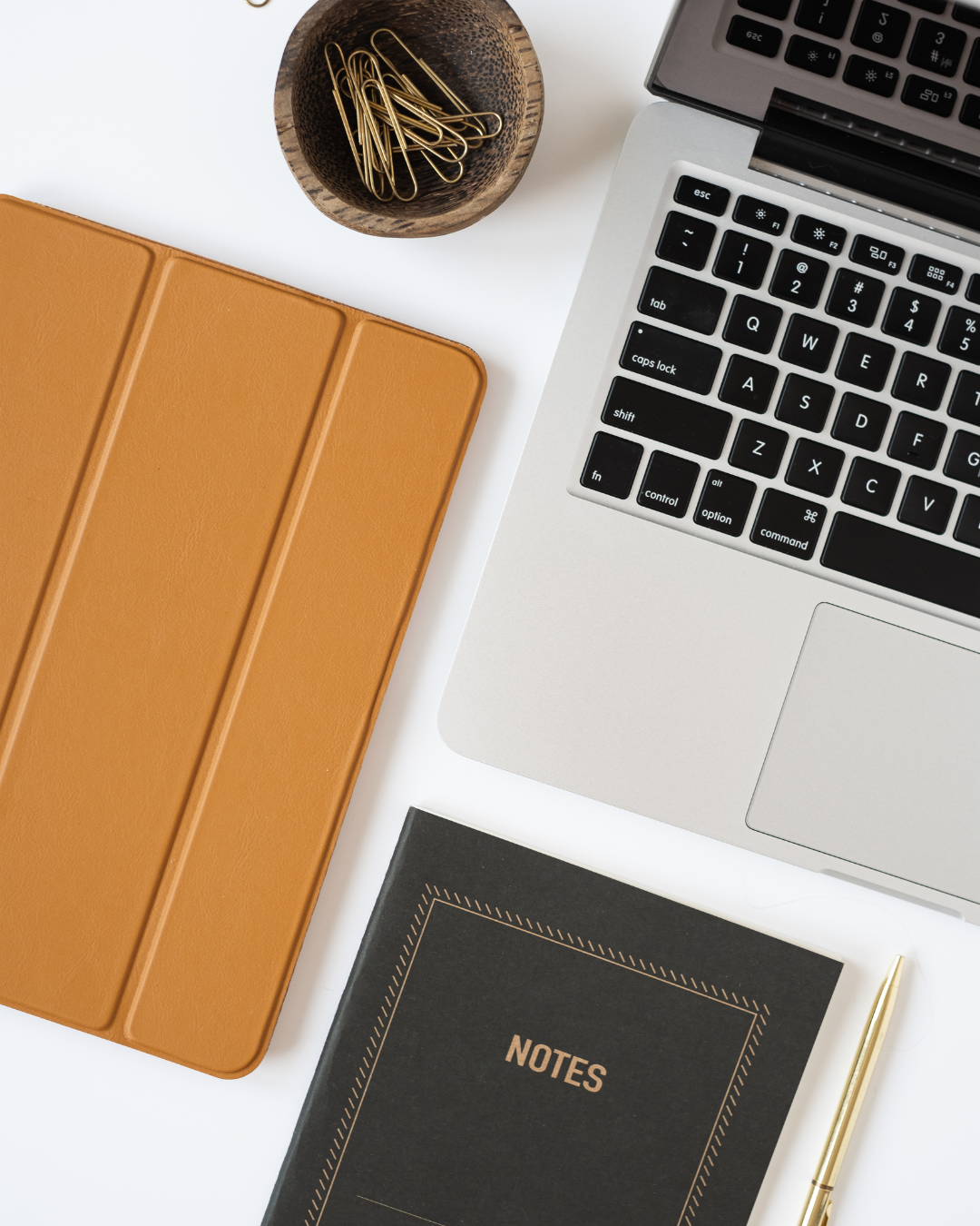 Workspace flat lay with laptop, black notebook, tan iPad cover, gold pen, and gold paperclips, representing The Uptown Collective's professional digital advertising environment