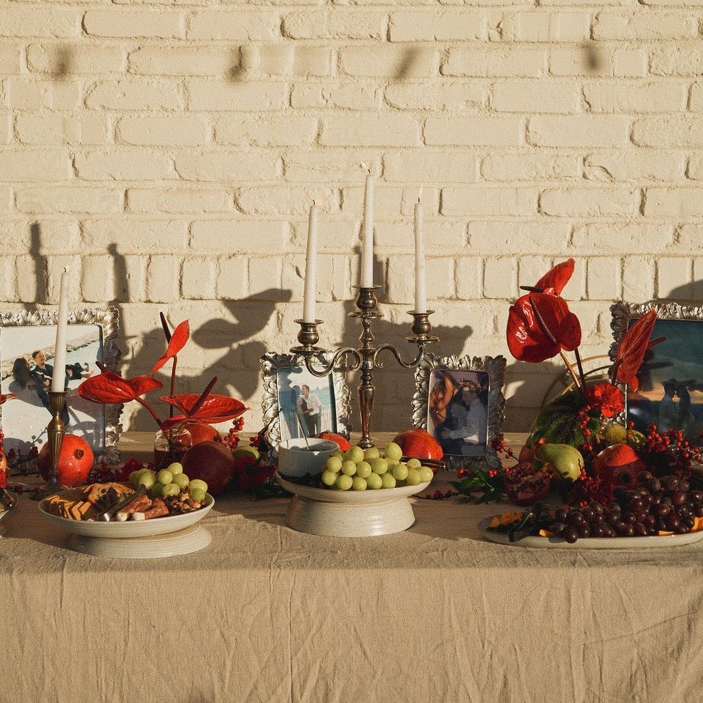 A decorated table with candles, framed photographs, and fruit including grapes, pomegranates, and apples, set against a white brick wall with shadows cast on it.