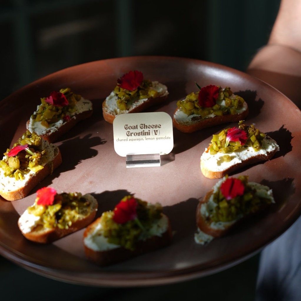A tray of goat cheese crostini topped with chard, asparagus, lemon gremolata, and garnished with pink edible flowers