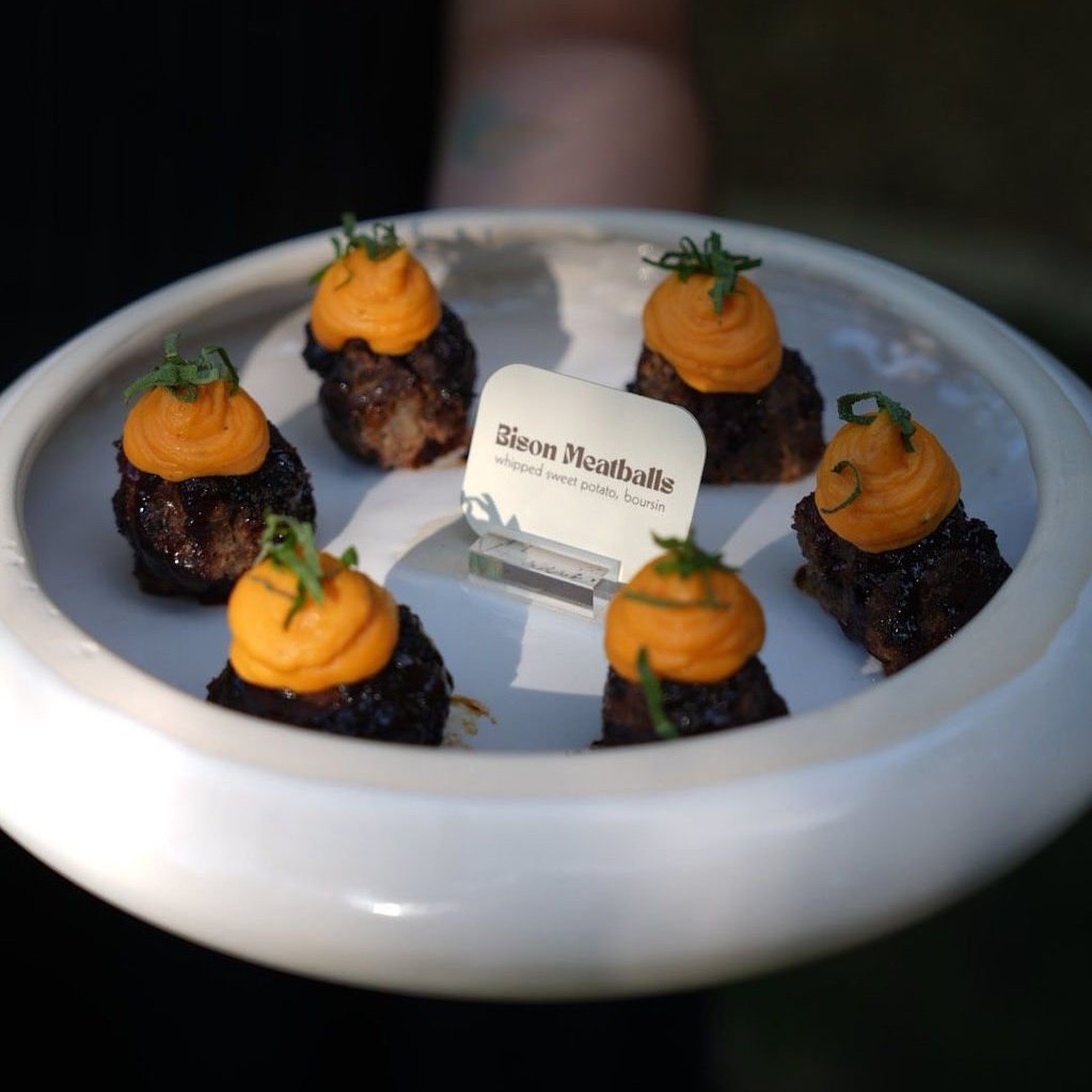 Plate of six Bison Meatballs topped with sweet potato mash and garnished with green herbs, on a white oval dish with a label reading 'Bison Meatballs, whipped sweet potato, boursin'