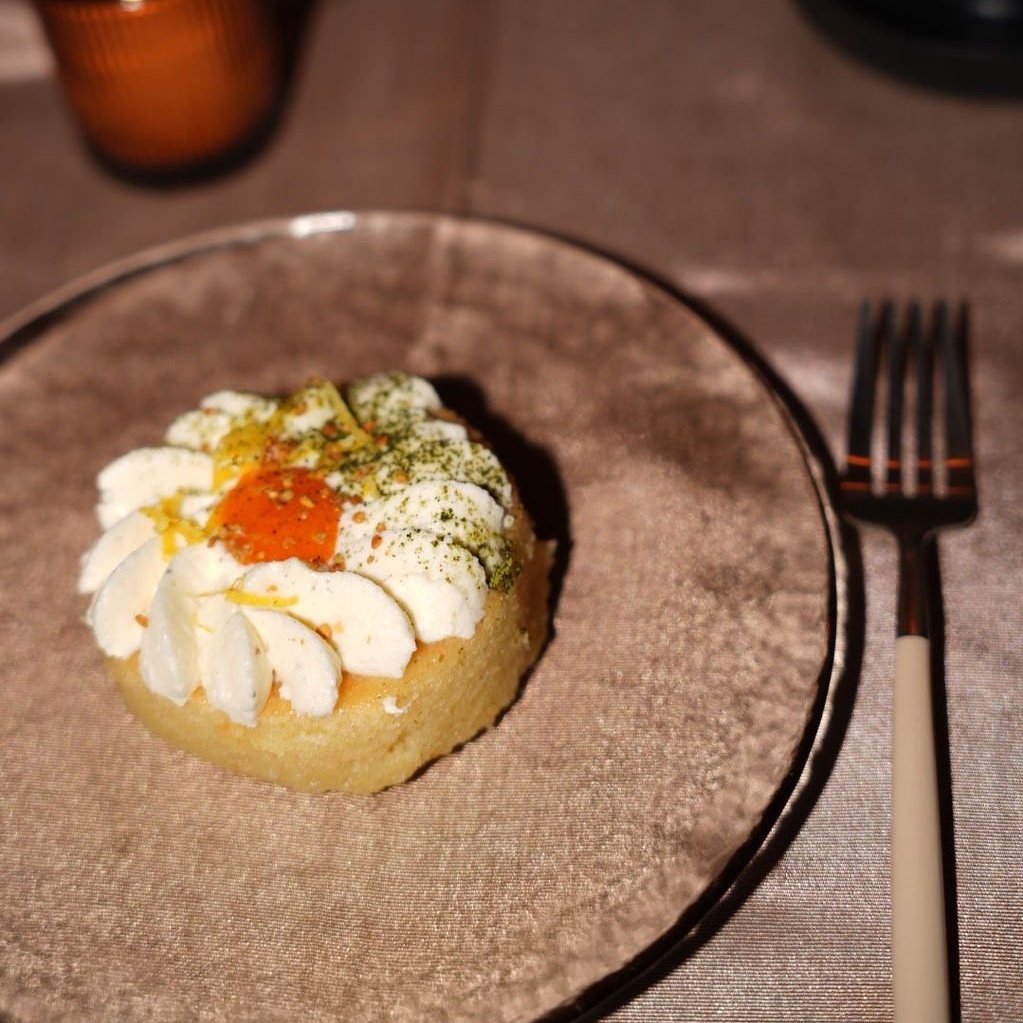 A small round cake topped with whipped cream, green herbs, a drizzle of orange syrup, on a transparent glass plate on a brown table with a fork nearby.