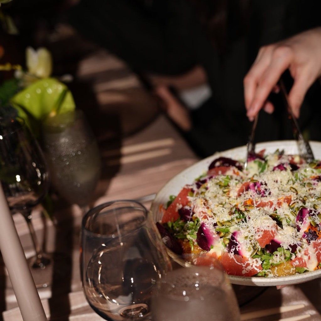 A person serving a colorful salad with greens, smoked salmon, purple cabbage, and grated cheese at a dinner table with glasses and a candle.