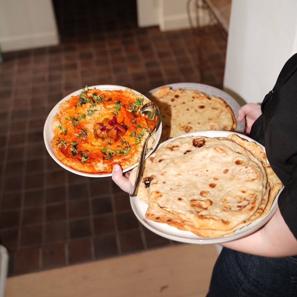 Person holding three plates of Indian cuisine, including naan bread, an onion pancake, and a bowl of vegetable curry garnished with cilantro and pomegranate seeds.