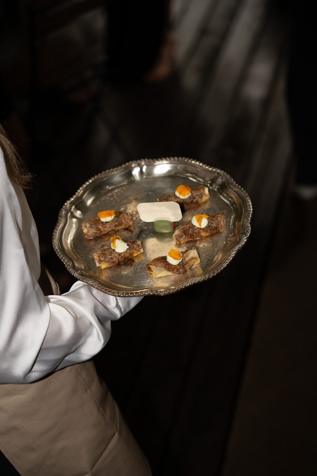 A server holding a silver tray with six small appetizers topped with cream, orange flower petals, and a rectangular piece of cheese.