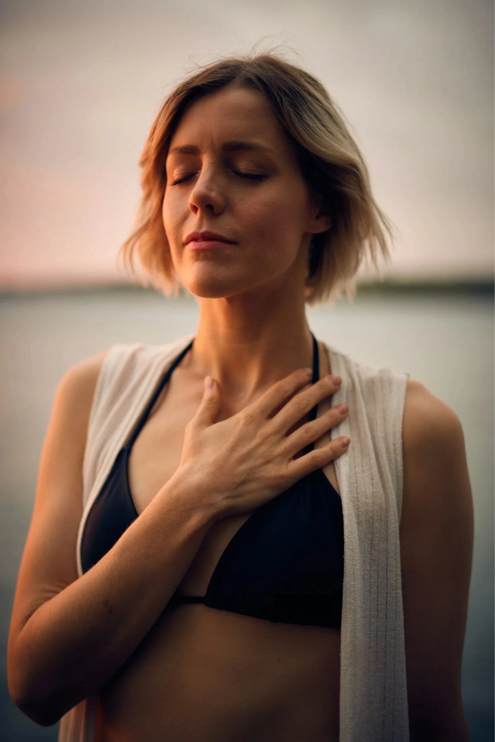 A woman stands by the sea, hand on her chest, breathing deeply as she meditates.