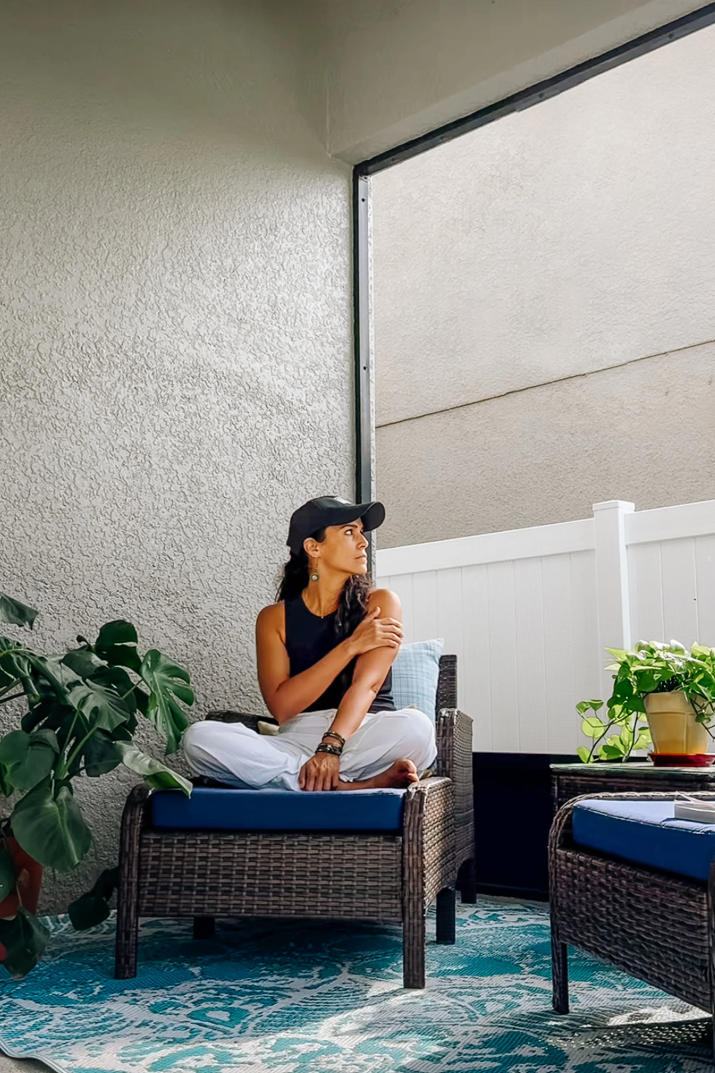 Sunshine Ritter sitting cross-legged on a wicker chair on a covered patio, breathing deeply. Potted plants and a white wooden fence are in the background.