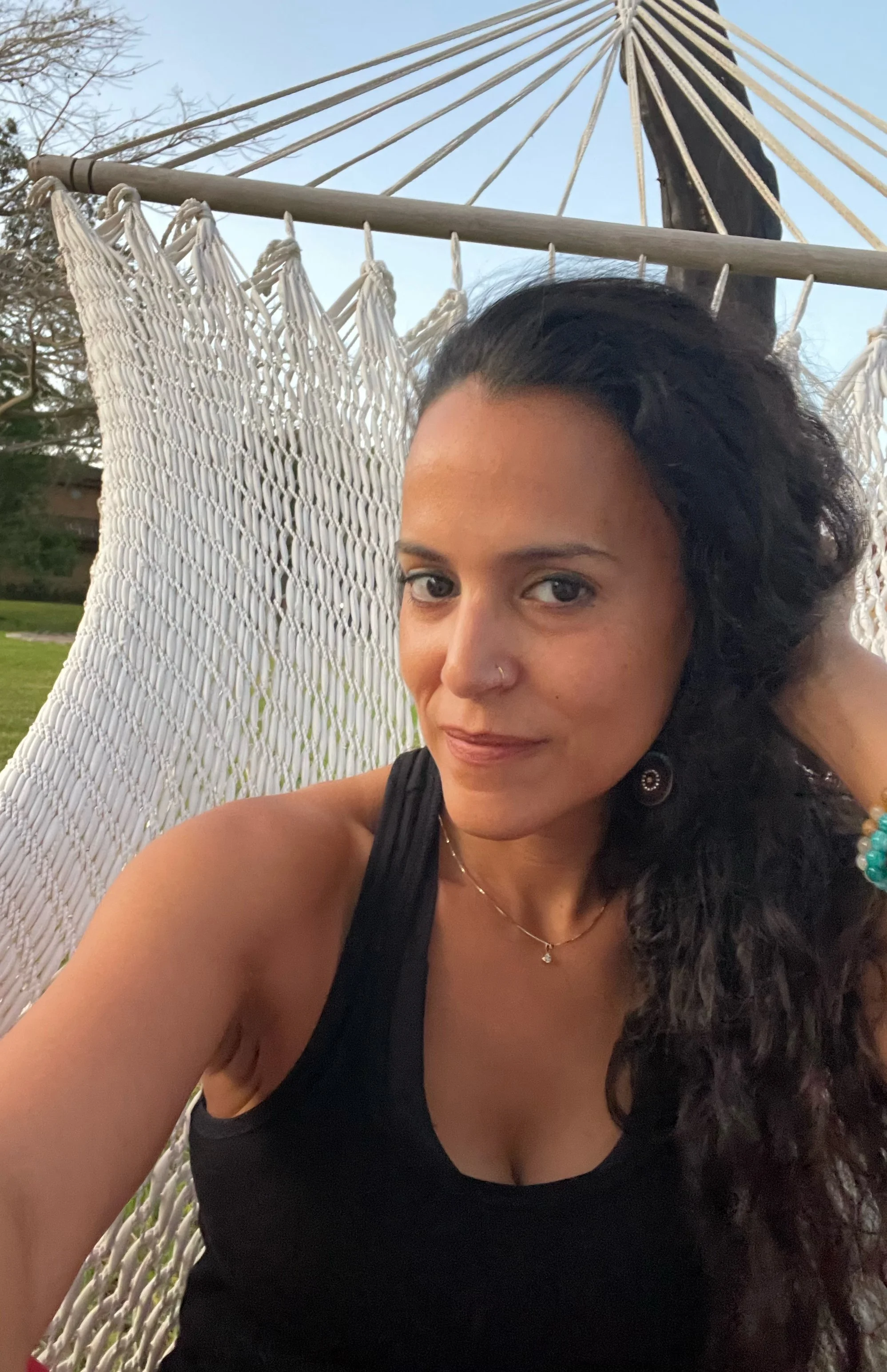 Sunshine wearing a black tank top and jewelry, sitting on a white rope hammock outdoors during daytime, with trees and a clear sky in the background.