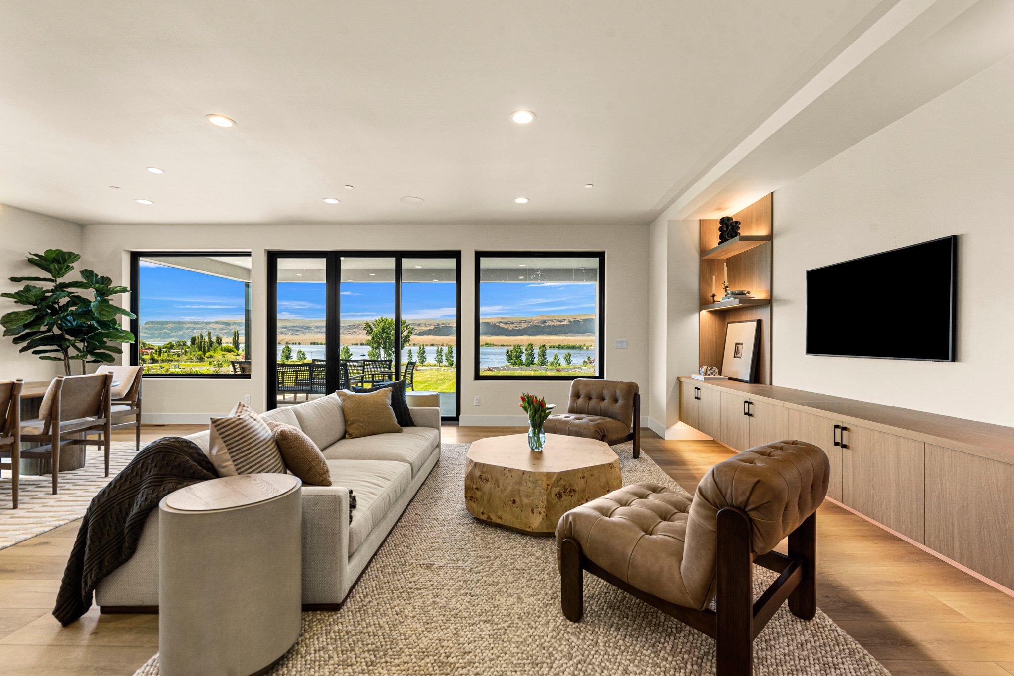Living room with large windows showing a scenic outdoor view of a lake, trees, and distant hills, furnished with beige sofas, brown leather chairs, a wooden coffee table, and decorative items, with a built-in media unit and a large flat-screen TV.