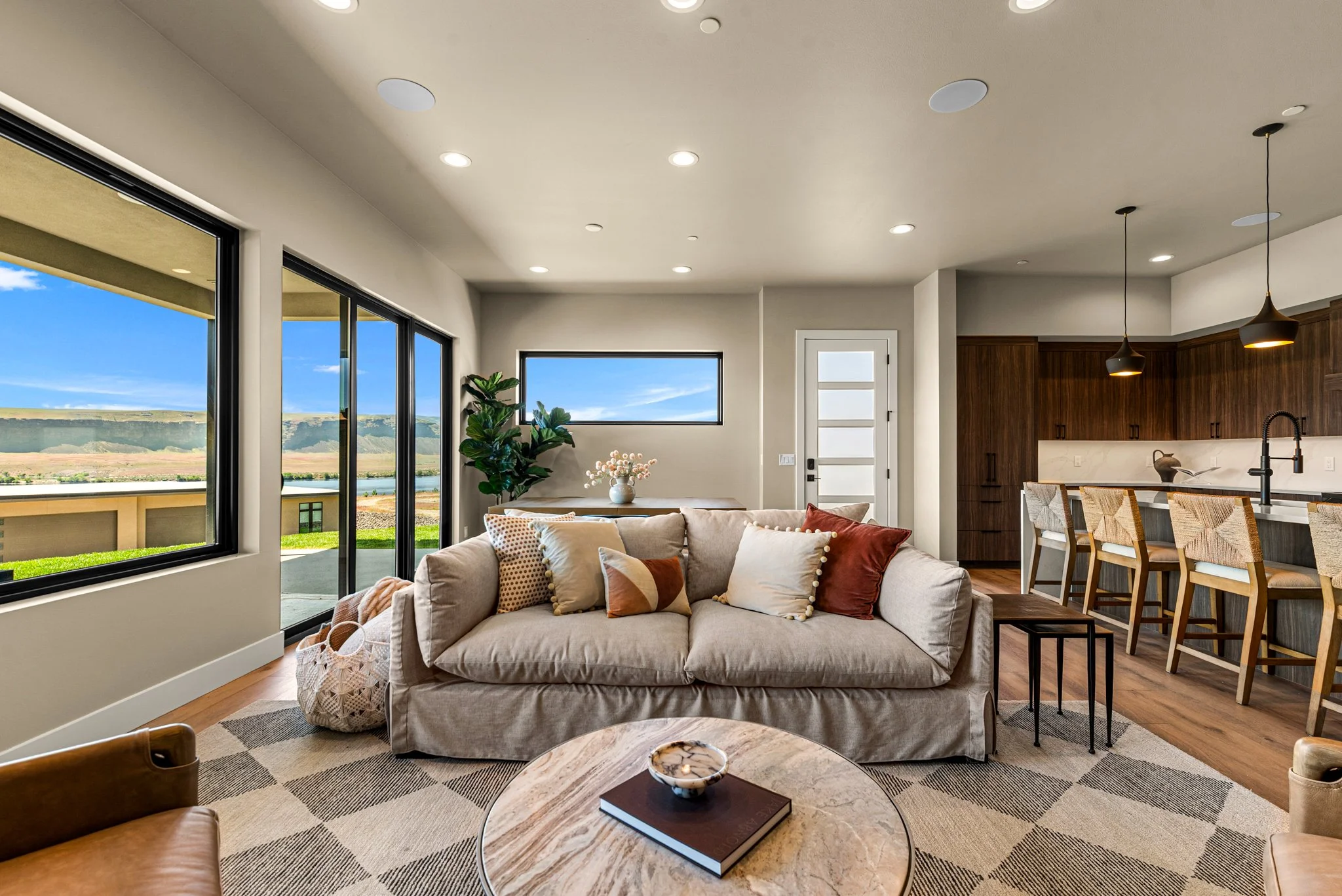 A modern, bright living room with large windows showing a scenic landscape outside. The room has a beige sofa with multiple decorative pillows, a round coffee table with a book and decorative bowl, and a patterned rug. The background features a kitchen area with wooden cabinets, hanging pendant lights, and a dining table surrounded by chairs.