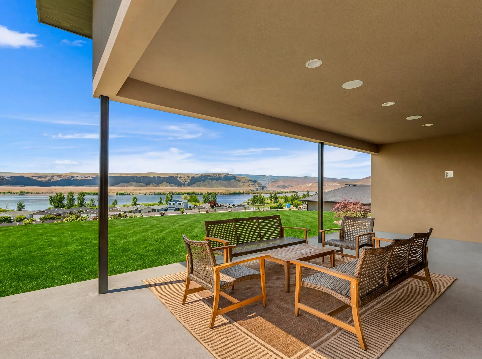 View of a patio with outdoor furniture, lush green lawn, trees, a body of water, hillside, and blue sky with clouds in the background.
