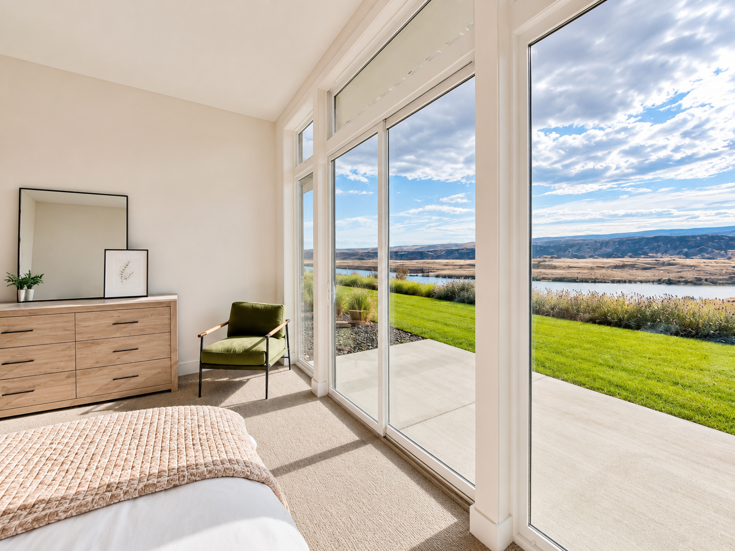 Room with large glass sliding doors showing a scenic lake and mountains outside, with a green lawn, bed partially shown, a wooden dresser with a mirror and framed artwork, a green armchair, and potted plants inside.