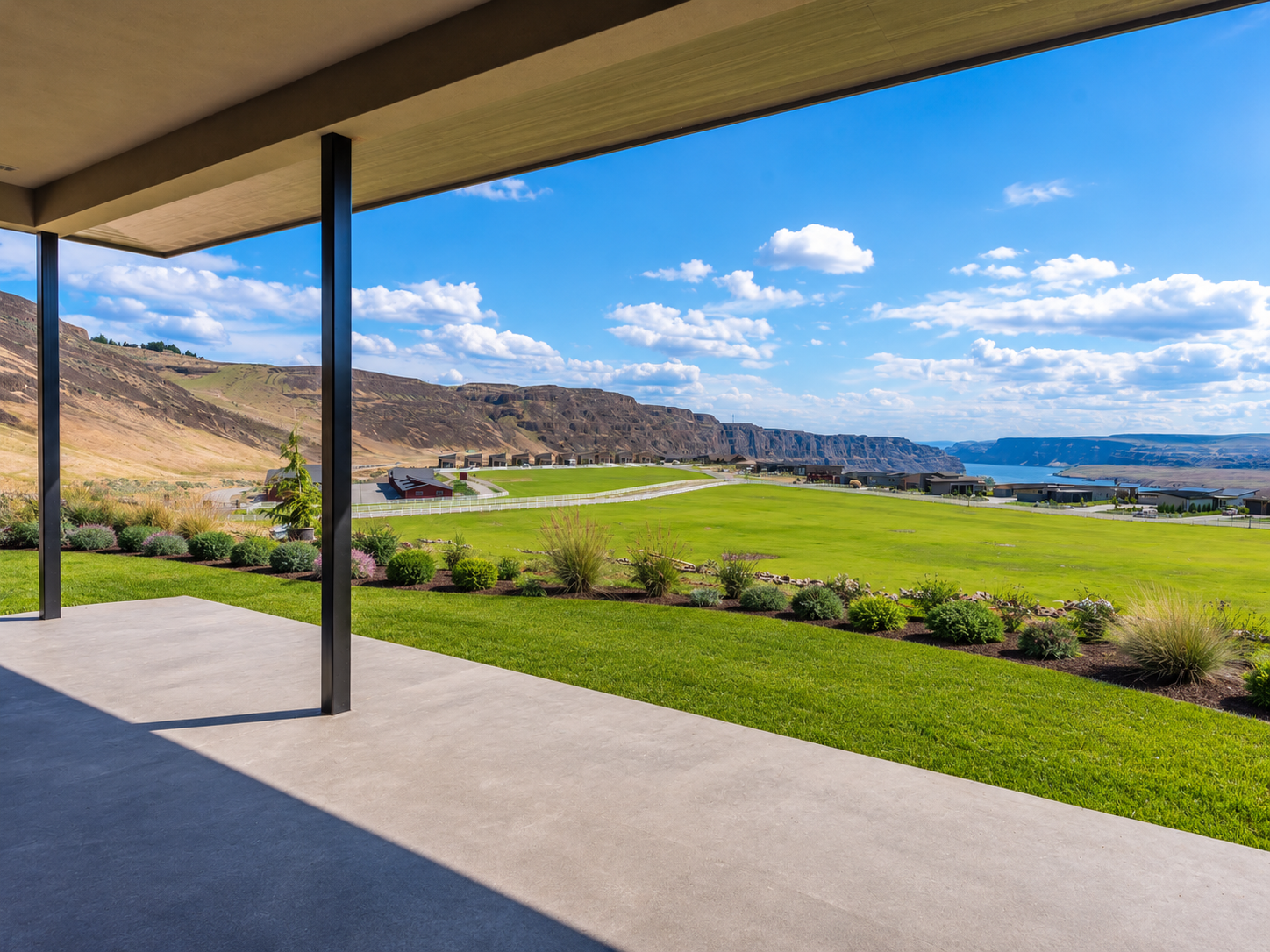 View from a covered patio overlooking a green field with landscaping, mountains, and a body of water in the distance under a blue sky with scattered clouds.