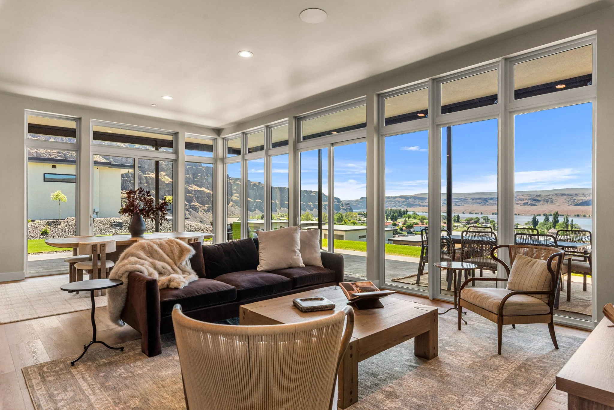 Living room with large windows showing mountains and a river view, furnished with a sofa, chairs, a wooden coffee table, and a dining table with chairs.
