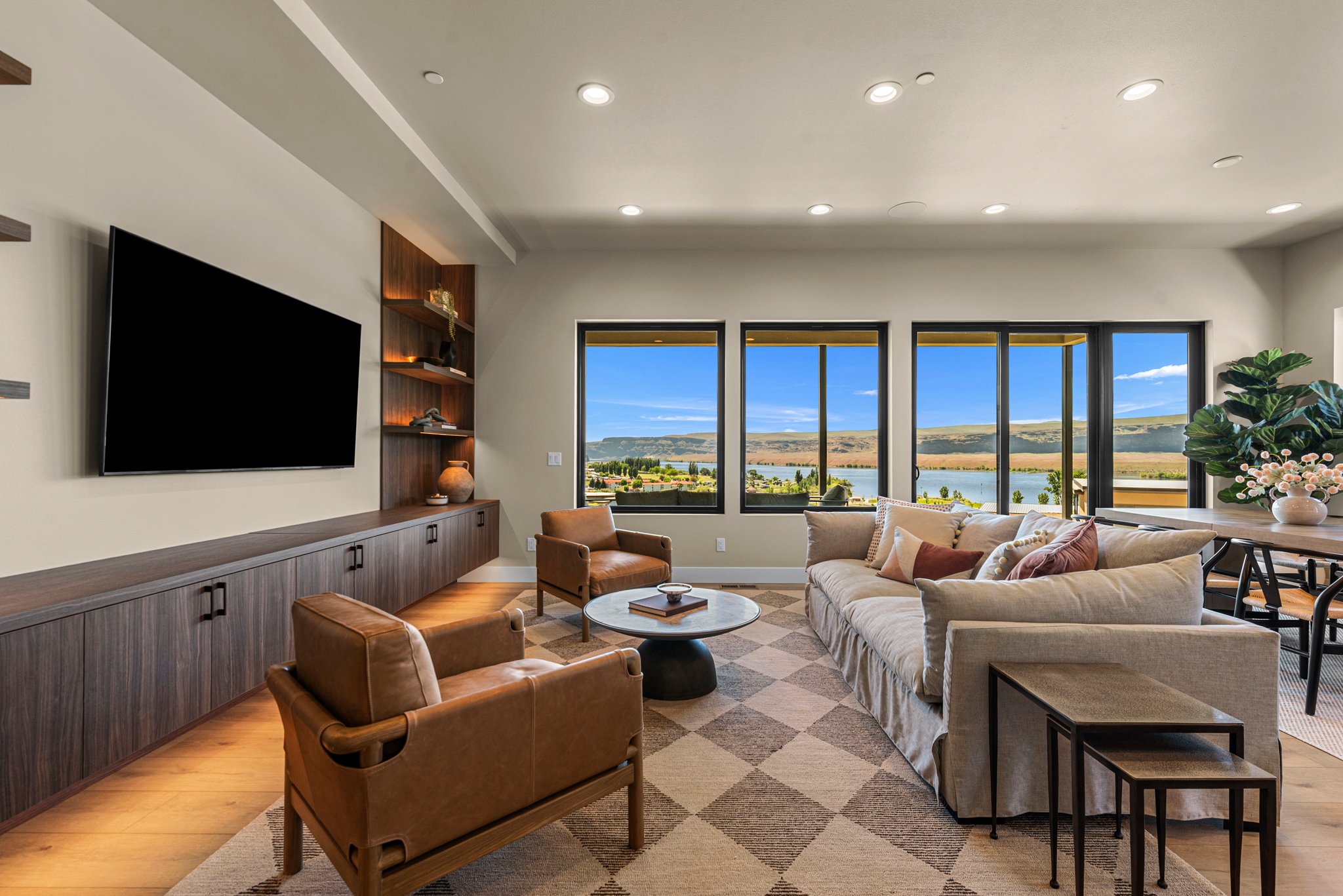 Modern living room with large windows showing a lake and hills outside, featuring a beige sofa, brown leather armchairs, a marble-topped coffee table, a built-in wooden media console with a flat-screen TV, and decorative items on shelves, complemented by indoor plants.