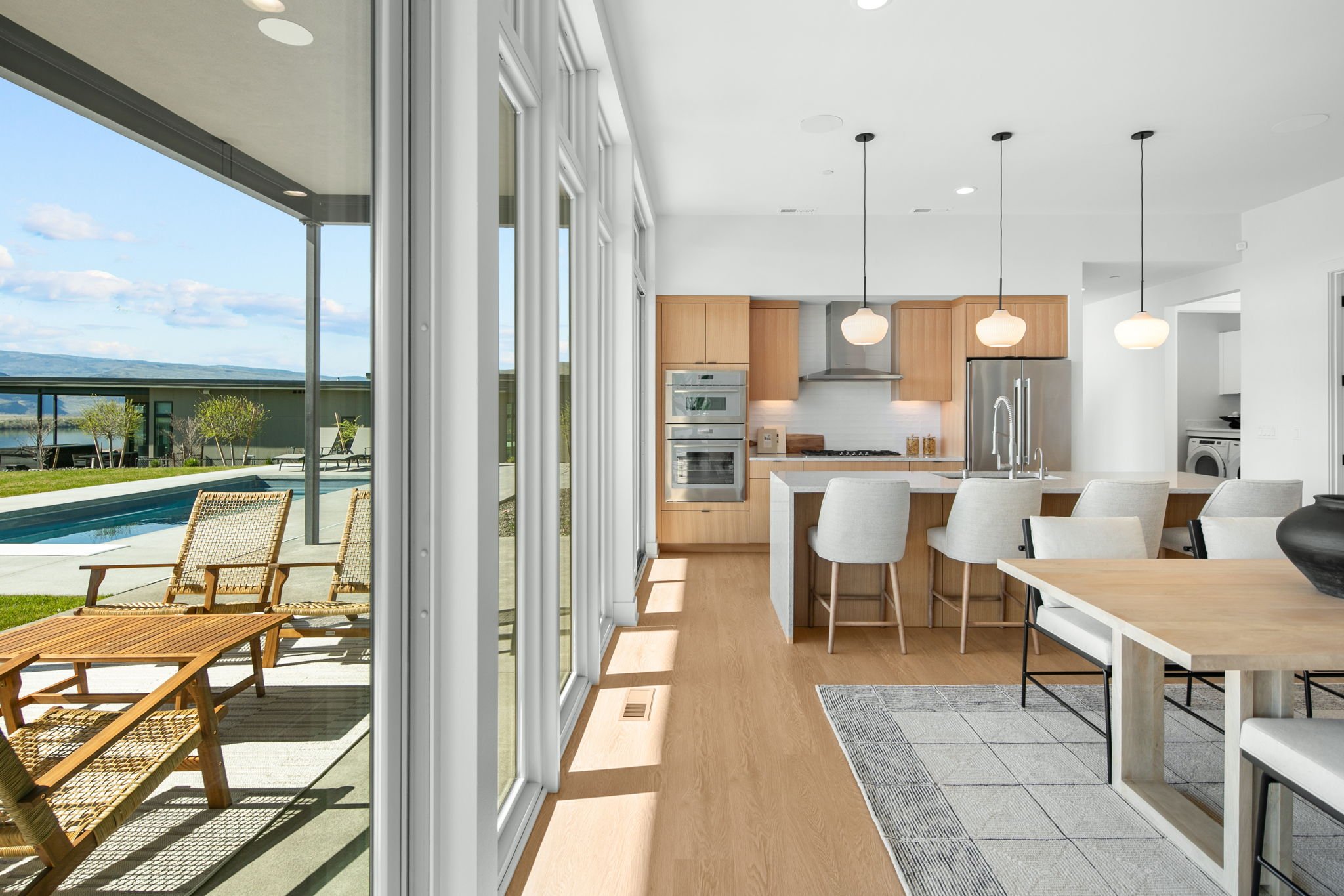 Modern open-concept kitchen and dining area with large sliding glass doors opening to a backyard with a swimming pool and lounge chairs, overlooking scenic hills under a partly cloudy sky.