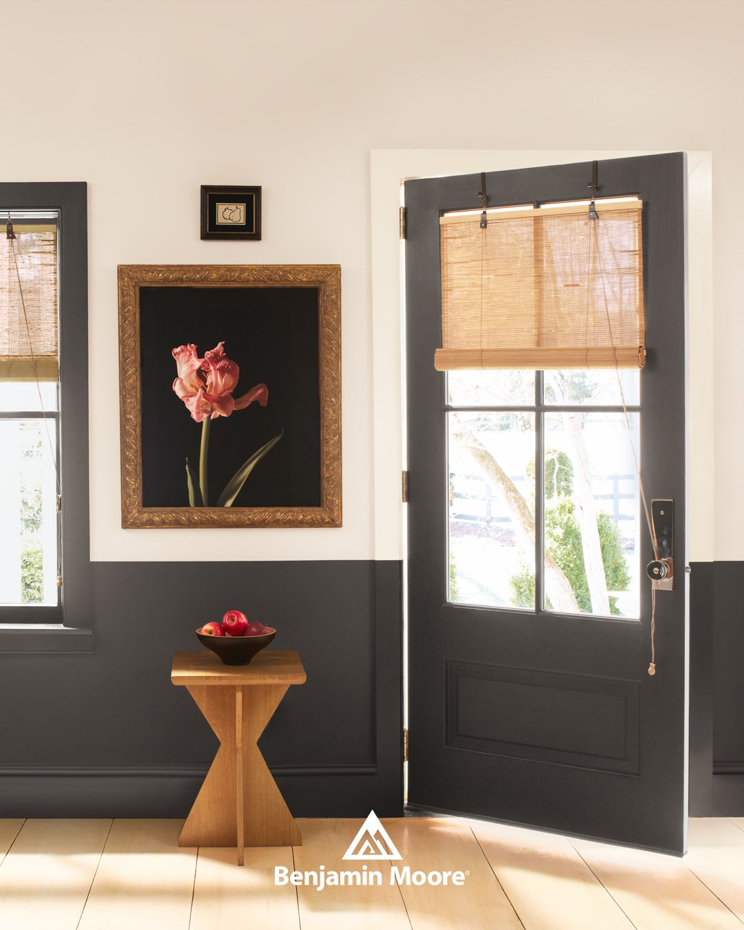 Interior entryway with a black door featuring a window with a bamboo shade, flanked by two windows with bamboo shades, a framed flower photograph on the wall, a small wooden side table with a bowl of red apples, and a small framed picture above the photograph, with natural light illuminating the space.