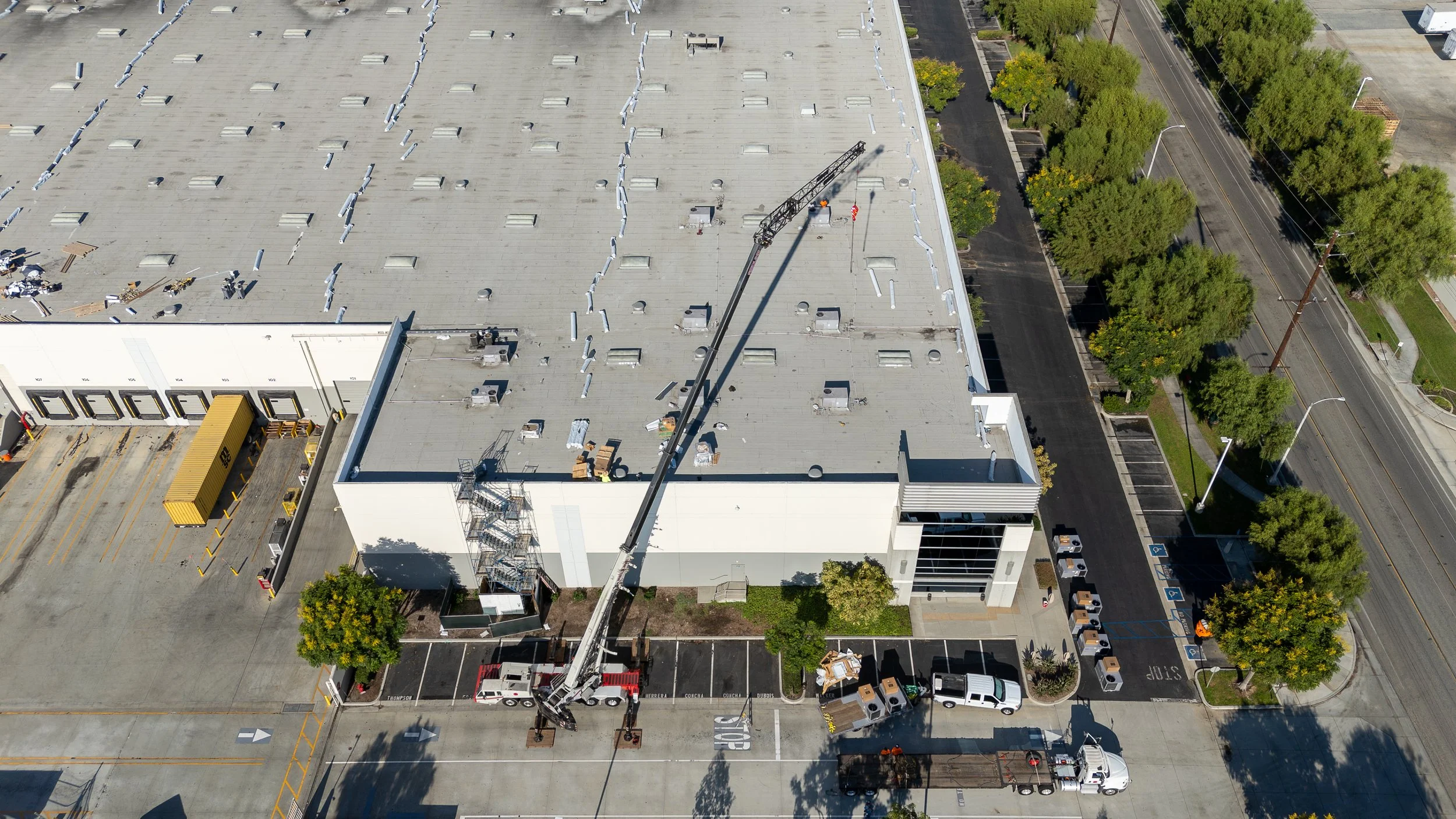 Aerial view of a large commercial building with a flat roof under construction, featuring a crane and construction materials, surrounded by parking lots and trees.