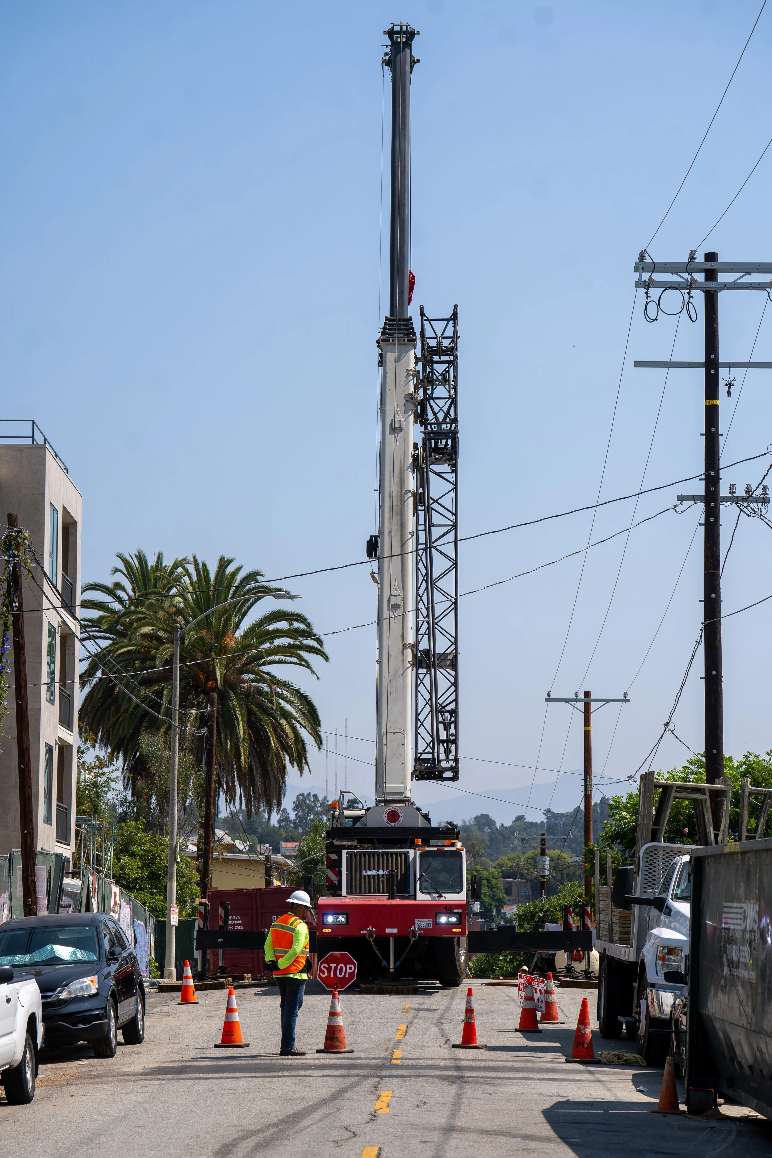 A construction worker in safety gear stands on a street near a large crane and utility poles with power lines, with cars parked along the road and traffic cones blocking the lane.