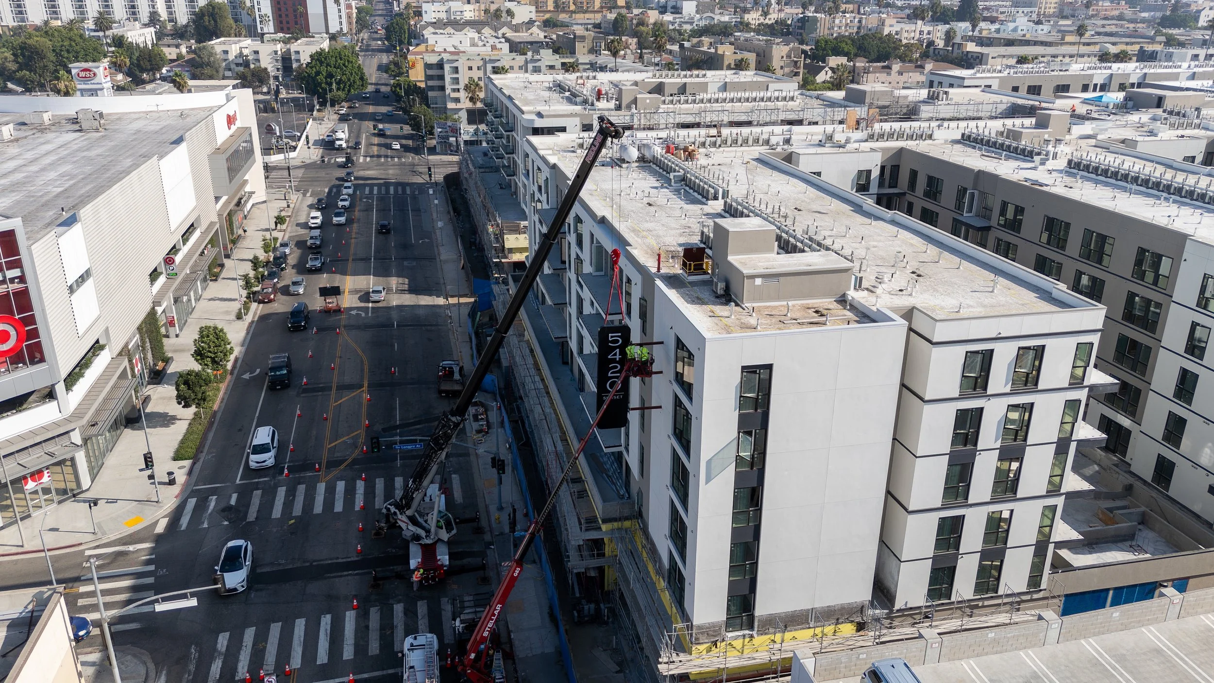 Construction workers operating cranes on the side of a white multi-story building under construction, located at a busy city intersection with cars and pedestrians.