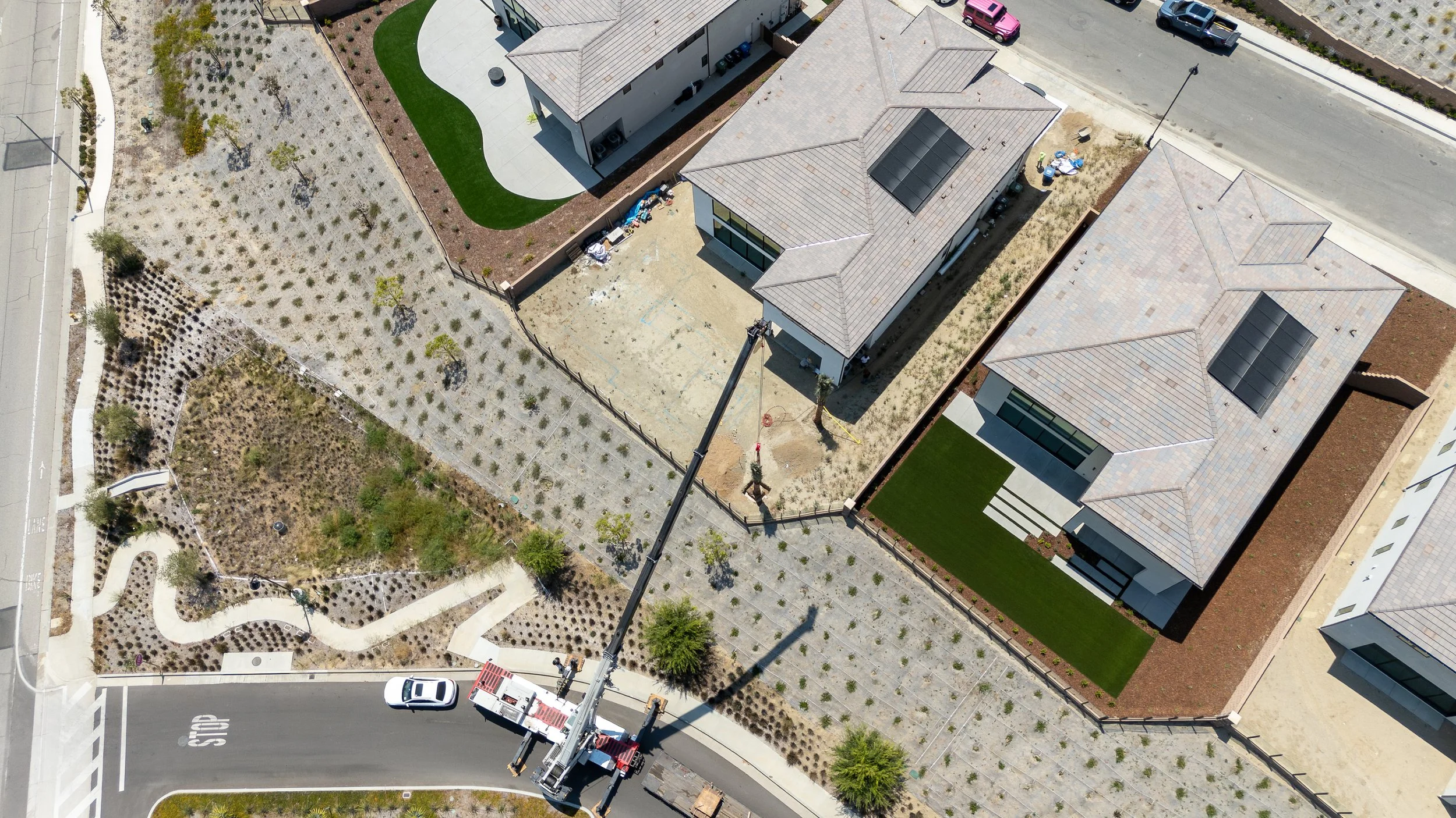 Aerial view of neighborhood with two new houses under construction, one with a green lawn, construction crane, and landscaping, with surrounding roads and parked cars.
