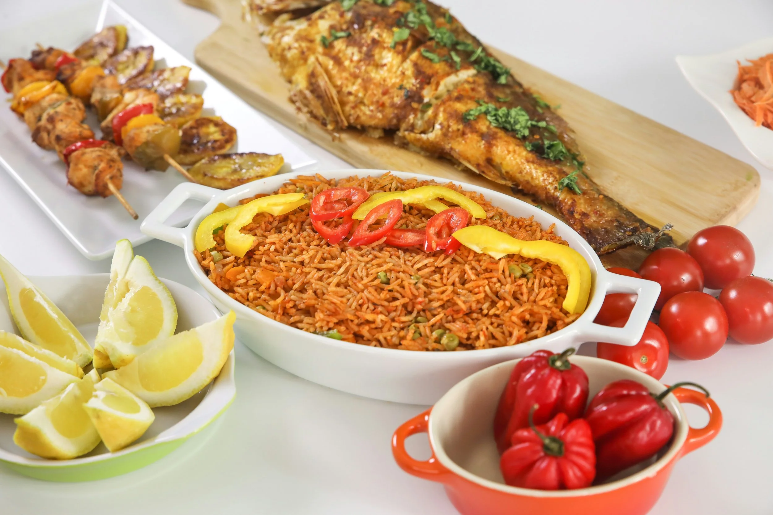 Assorted dishes including skewered grilled meat, baked fish garnished with herbs, spiced rice with sliced peppers, cherry tomatoes, lemon wedges, and fresh red tomatoes.