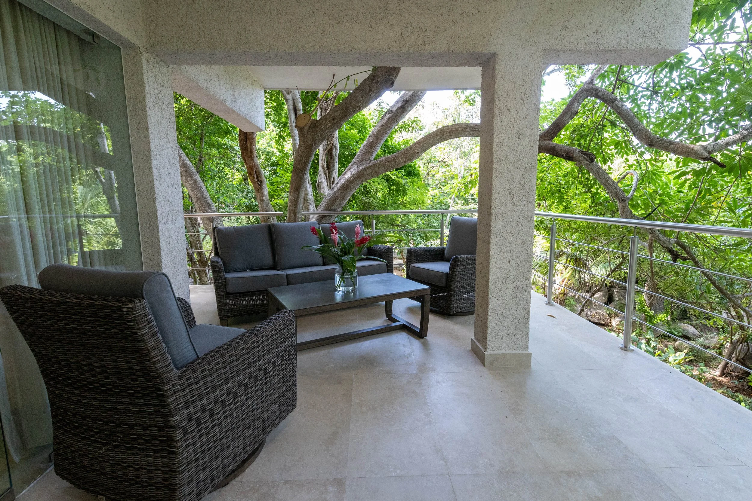 Covered outdoor patio with wicker seating, a coffee table, and tropical flowers, overlooking a lush green forest with trees surrounding the balcony railing.