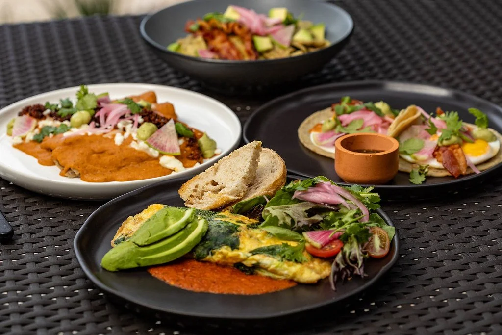 Assorted gourmet breakfast plates featuring omelets, tacos, fresh avocado, pickled onions, and vibrant sauces served on a woven outdoor dining table.