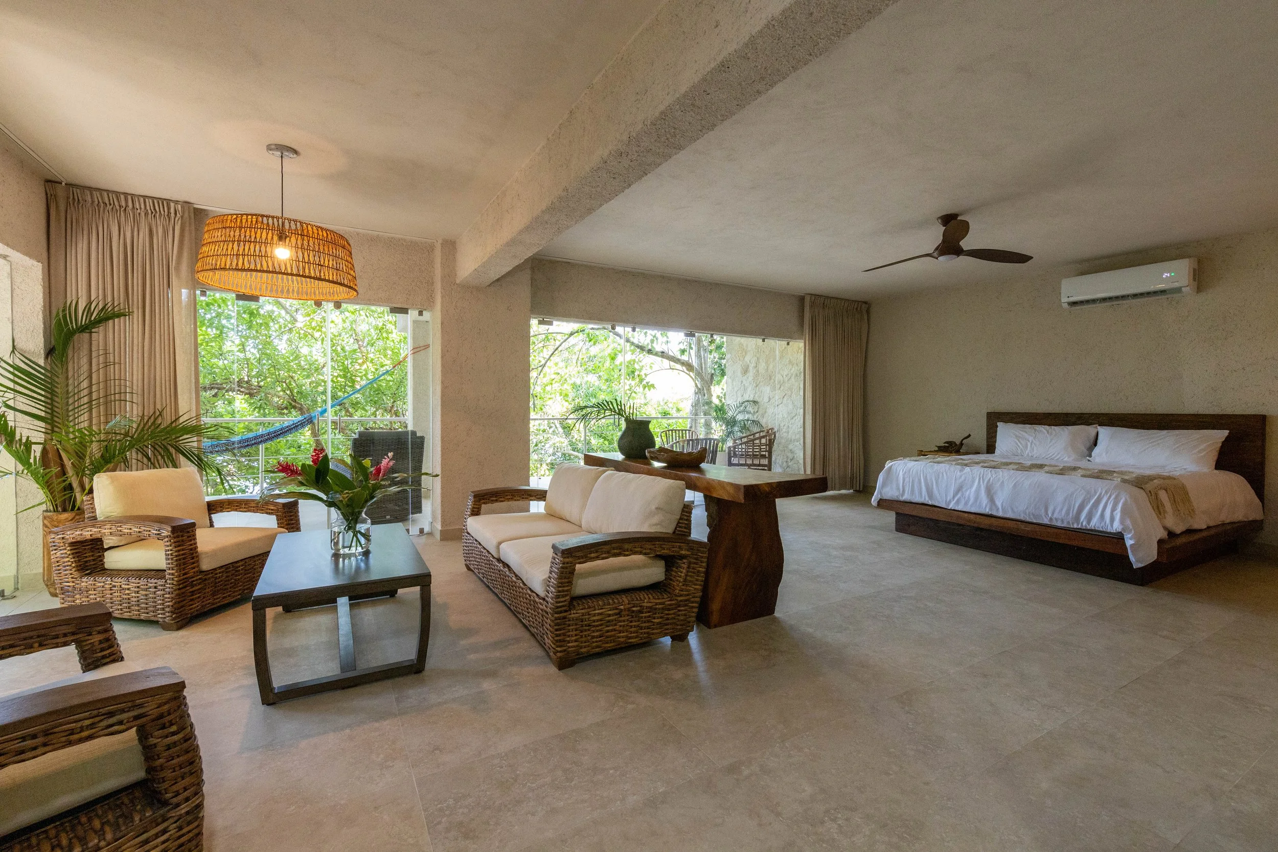Spacious open-concept suite with wicker seating, a dining table, and a king bed, featuring natural textures, large windows, and views of lush tropical greenery.