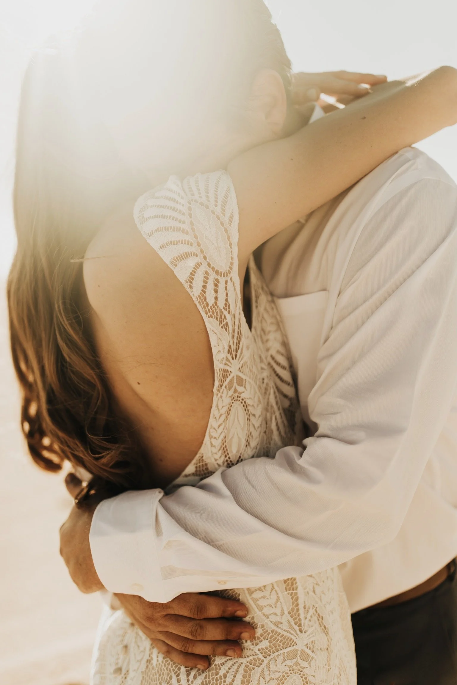 Close-up of a couple embracing during a beach wedding, with soft sunlight creating a warm, intimate glow and lace details visible on the bride’s dress.