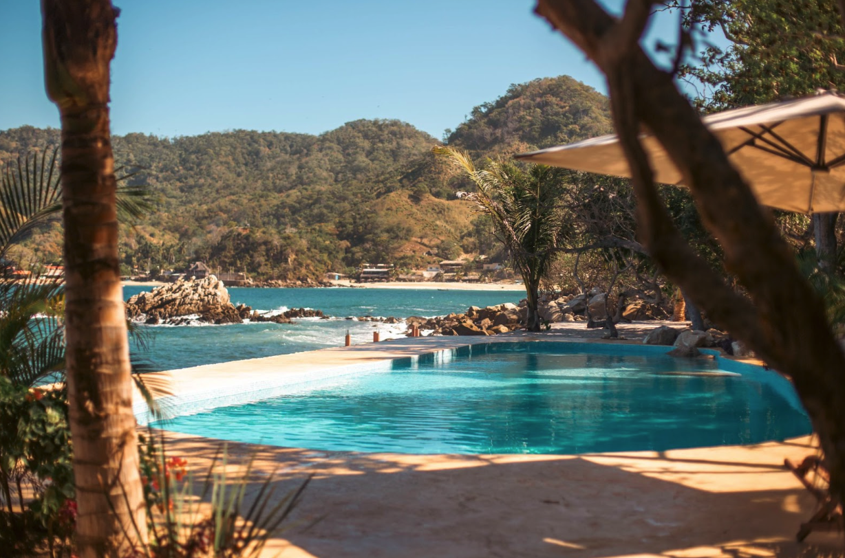 Infinity pool overlooking a turquoise ocean with rocky coastline and lush green hills in the background, framed by palm trees and shade from an umbrella.