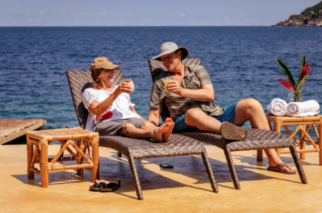 Older couple relaxing on lounge chairs by the ocean, smiling and sharing tropical drinks with towels and flowers on a small table beside them.