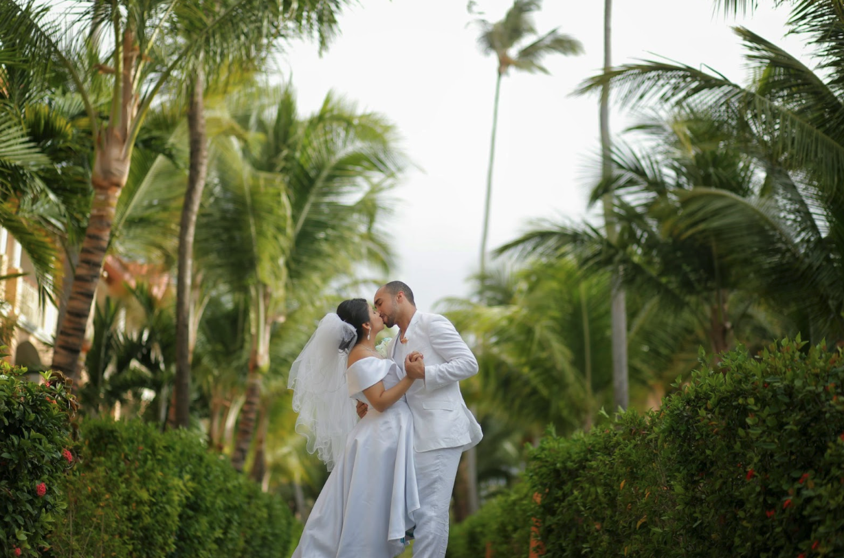 Bride and groom sharing a kiss in a tropical garden pathway surrounded by lush palm trees and greenery during their outdoor wedding.
