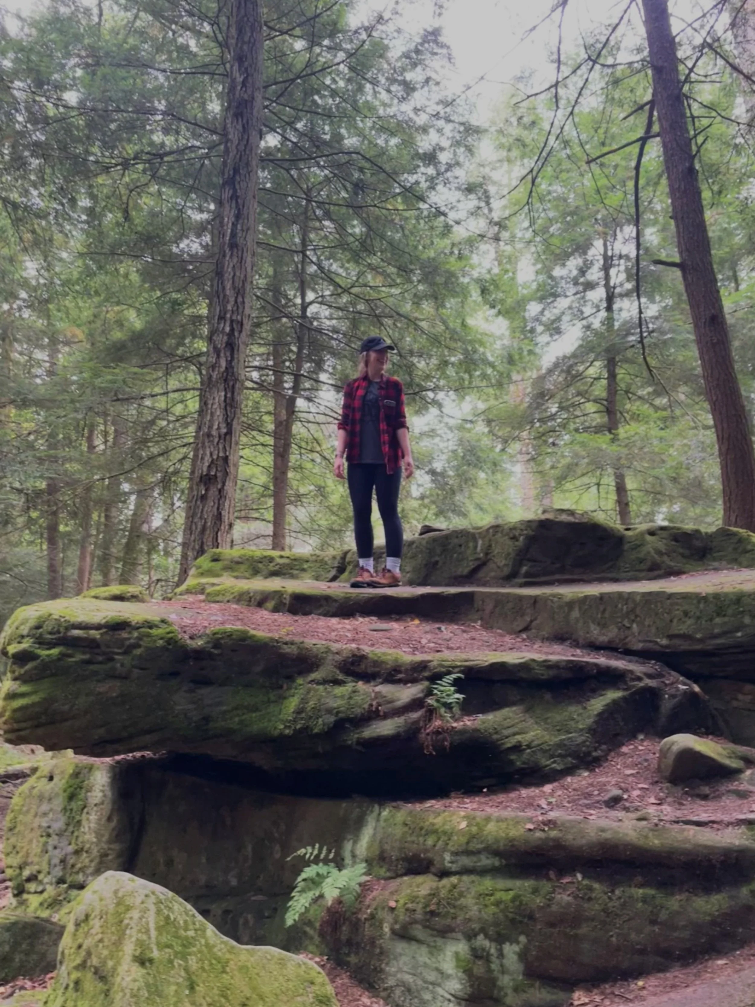 A woman standing on a mossy rock formation in a dense forest, surrounded by tall trees and greenery.
