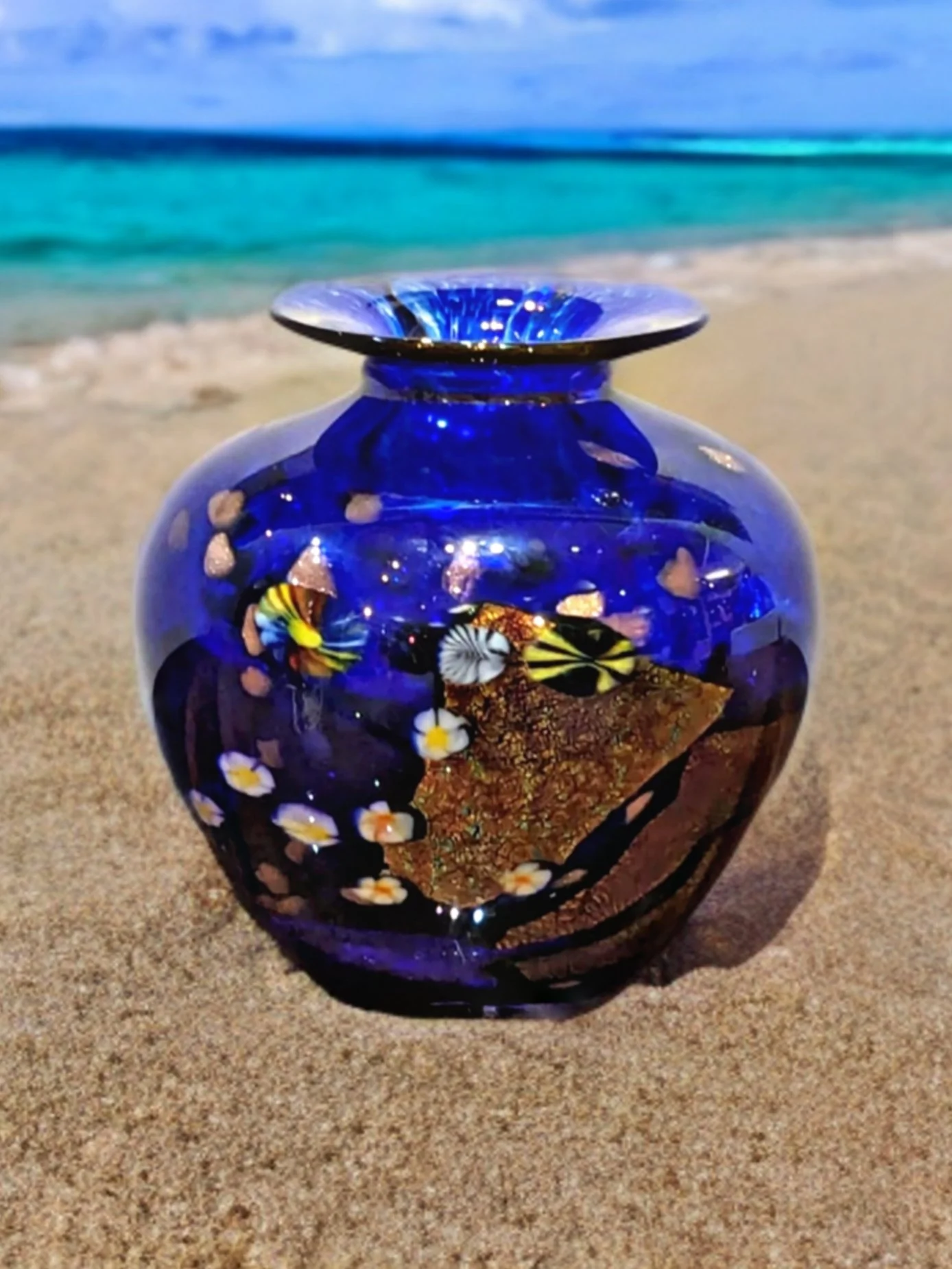 A vibrant blue glass vase with floral and beach-themed design on a sandy beach with ocean and sky in the background.