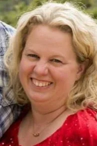 Close-up of a smiling woman with curly blonde hair, wearing a red top, outdoors with greenery in the background.