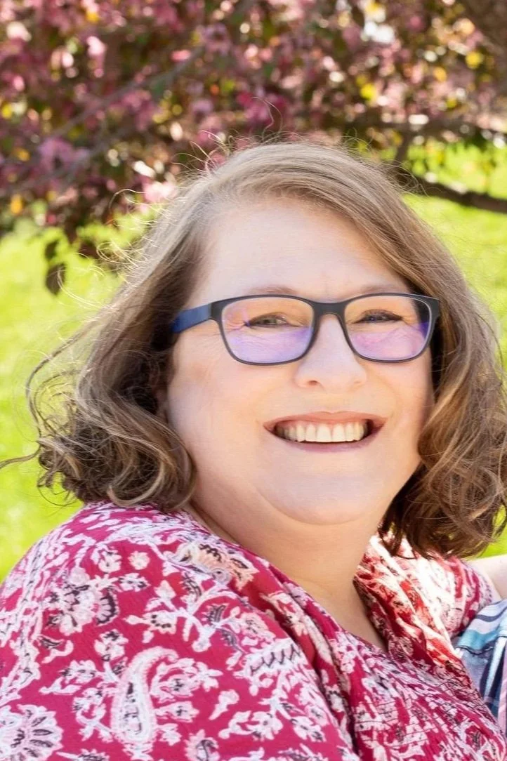 A woman with shoulder-length curly brown hair, wearing glasses with purple-tinted lenses, smiling outdoors in front of a pink flowering tree.