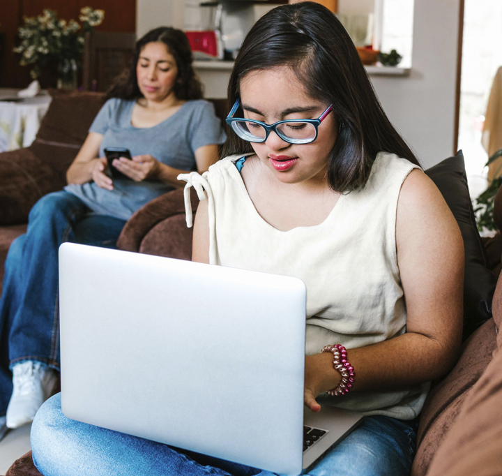 A young woman with glasses works on her laptop at home while a family member relaxes nearby.