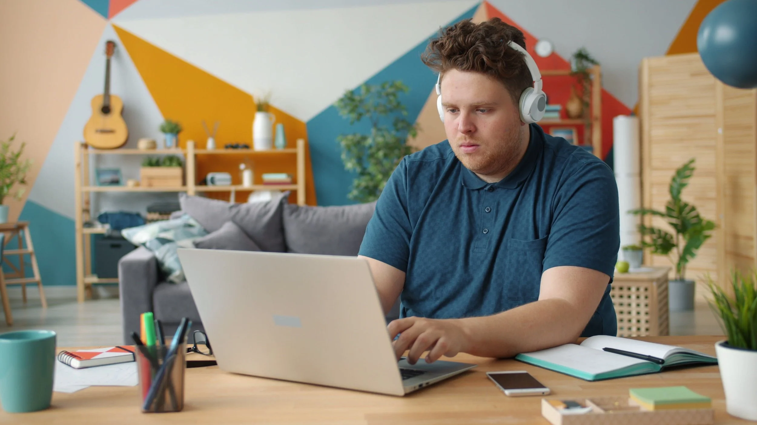 A young man wearing noise-canceling headphones works on his laptop at a desk in a bright, colorful home office.