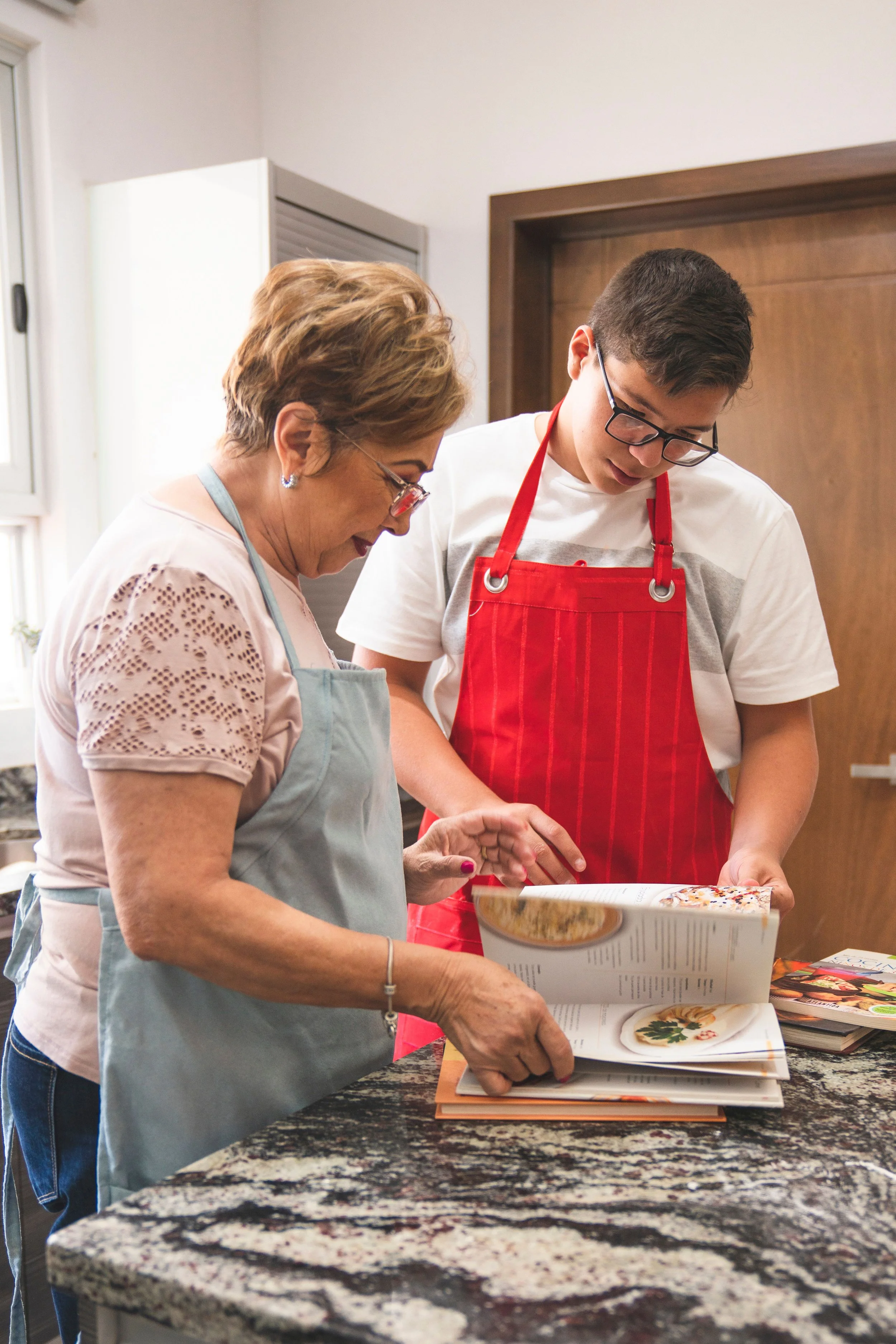 A young man in an apron follows a recipe in the kitchen as his grandmother reads along beside him.