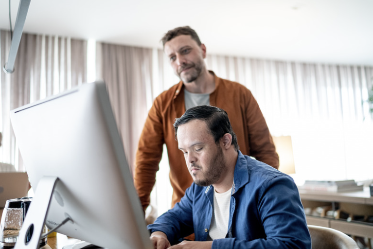 A man works at a computer while his father stands behind him, offering guidance.