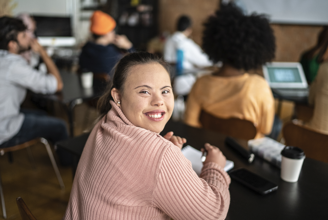 A young woman smiles over her shoulder while taking notes at a table in a busy workshop or class.