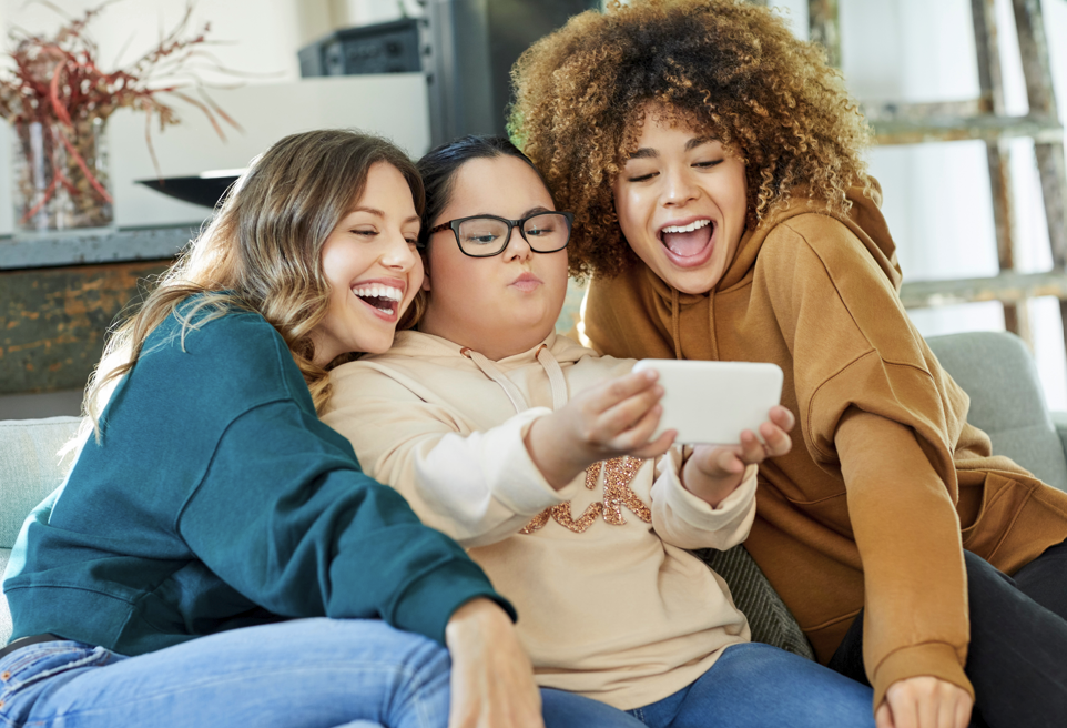 Three young adult friends laugh together on a couch while taking a selfie.